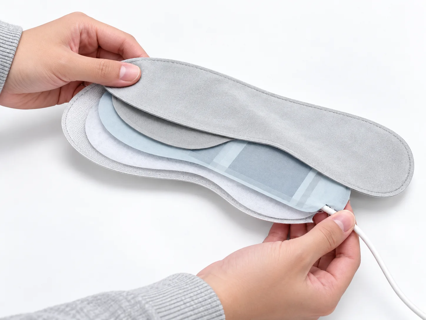 Hands holding a layered eye mask with its heating layer and cable visible